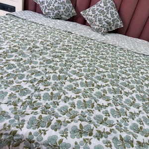 Könnte beinhalten: Ein weißer Bettüberwurf mit grünem Blumenmuster. Der Bettüberwurf liegt auf einem Bett mit zwei passenden Kissen mit Blumenmuster.