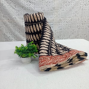 May include: A rolled-up black and beige patterned textile with a red border, next to a small potted plant. The textile features a repeating design of black shapes on a beige background. The border has a red and beige pattern.