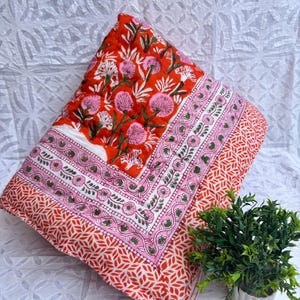 May include: A quilted throw blanket with a floral pattern in shades of red, pink, and white. The blanket features a detailed border design and a repeating leaf pattern on the edges. The blanket is folded and placed on a white textured surface.