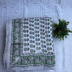 Puede incluir: Un edredón floral blanco y verde doblado con un borde verde con estampado de cachemira. El edredón presenta un patrón repetitivo de flores verdes y marrones sobre un fondo blanco. Una pequeña planta verde se coloca a un lado.
