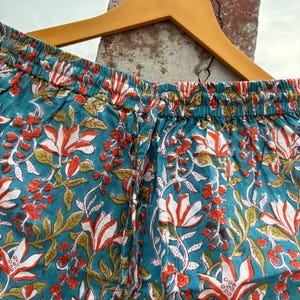K&ouml;nnte beinhalten: Tealblaue Baumwollhose mit wei&szlig;em und orangefarbenem Blumendruck. Die Hose hat einen elastischen Bund und einen Kordelzug.