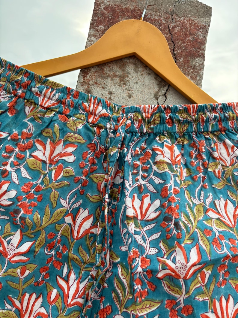K&ouml;nnte beinhalten: Tealblaue Baumwollhose mit wei&szlig;em und orangefarbenem Blumendruck. Die Hose hat einen elastischen Bund und einen Kordelzug.