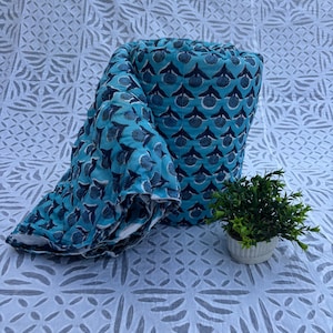 May include: A rolled-up, turquoise quilt with a repeating floral pattern in navy blue. The quilt is partially unrolled, revealing the white backing. A small potted plant sits nearby on a white patterned surface.