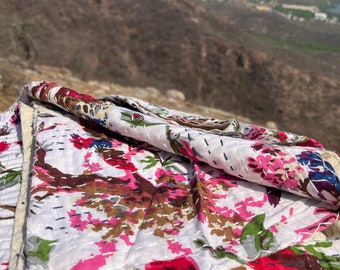 May include: A white kantha quilt with a floral pattern in shades of pink, red, green, and brown. The quilt is folded and lying on a rocky surface with a mountain range in the background.