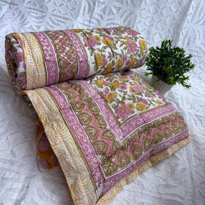 Könnte beinhalten: Eine florale Steppdecke, teilweise aufgerollt. Sie hat einen weißen Hintergrund mit rosa und gelben Blumen, grünen Akzenten und einem rosa Rand. Die Ränder sind hellgelb mit einem wellenförmigen Muster.