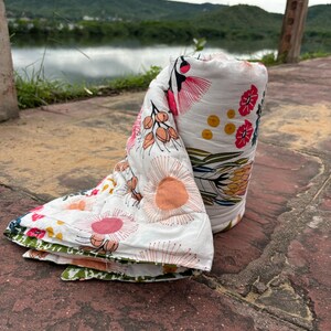 May include: A white muslin swaddle blanket with a floral print featuring pink, orange, and yellow flowers. The blanket is rolled up and lying on a brown tiled surface.