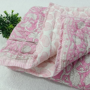 May include: A pink and white cotton quilt with a floral print. The quilt is folded in half and has a white background with pink flowers and leaves. The quilt is made of a soft, lightweight fabric and is perfect for keeping warm on a cool night.