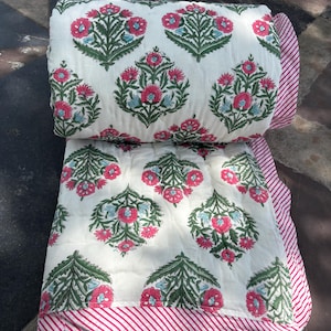 May include: Rolled-up white quilt with a floral pattern in pink, green, and blue. The quilt's edge features a pink and white striped border. The floral design is repeated across the quilt's surface.
