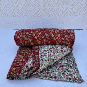 Op de afbeelding: Een rood en wit bloemmotief quilt met een gestikt ontwerp. De quilt is dubbelgevouwen en opgerold.