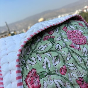 May include: A green and white floral patterned quilt with pink flowers and a white border with pink stripes. The quilt is folded in half and the floral side is facing up.