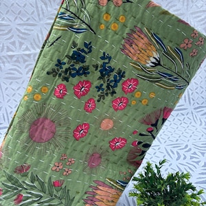 May include: A folded green quilt with a floral pattern. The quilt features large pink, orange, and yellow flowers, along with smaller pink and blue floral accents. The quilt is hand-stitched with white thread.