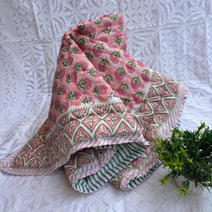Puede incluir: Un edredón floral rosa doblado con un borde geométrico blanco y verde. El edredón presenta un patrón repetitivo de flores rosas sobre un fondo rosa claro. El borde tiene un diseño geométrico con acentos verdes.