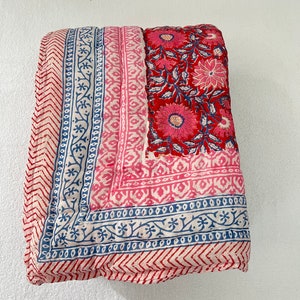 May include: A pink and red cotton quilt with a floral pattern and blue and white border. The quilt is folded in half and has a chevron pattern on the bottom edge.