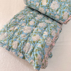 Op de afbeelding: Een blauwe en witte katoenen deken met bloemenprint en een roze rand. De deken is dubbelgevouwen en ligt op een wit oppervlak.