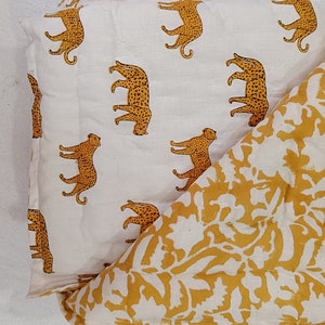 May include: A reversible quilted blanket featuring a white side with a repeating pattern of orange leopard print and a yellow side with a white floral design. The blanket is folded, showcasing both patterns.