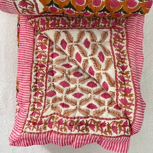 May include: A white and pink cotton quilt with a floral pattern. The quilt is folded in half and has a pink and white striped border.