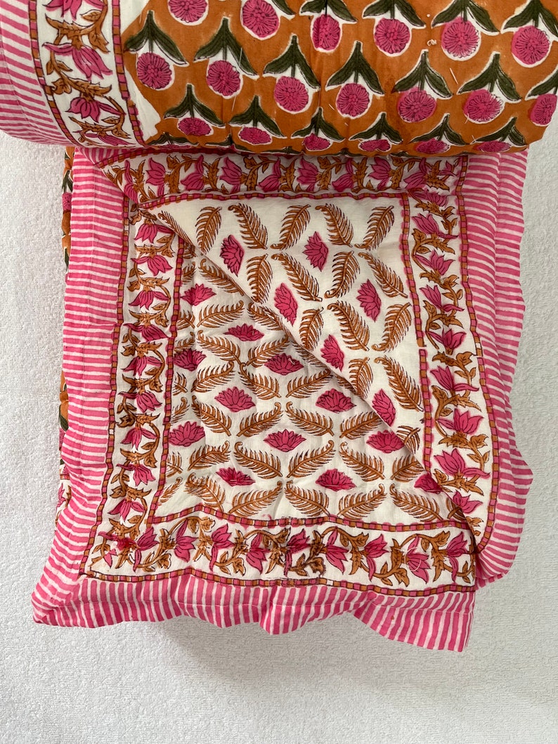 May include: A white and pink cotton quilt with a floral pattern. The quilt is folded in half and has a pink and white striped border.