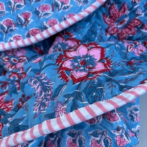 May include: A blue and pink floral fabric with a white and pink striped border. The fabric is quilted and has a soft, textured feel.