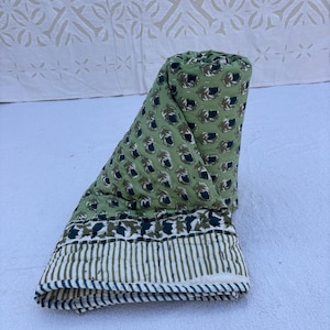 May include: Rolled-up green quilt with a repeating pattern of dark green and black leaf-like shapes. The quilt's border features vertical stripes in green and white. The background is a white surface.