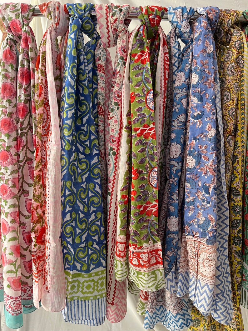 May include: A collection of colorful cotton scarves with various floral and geometric patterns. The scarves are hanging on a rack and are ready to be worn.