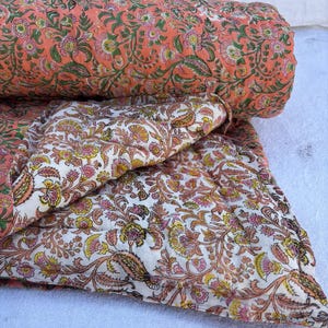 以下が含まれることがあります： ピンクと白の花柄の綿のキルトで、ペイズリー柄が施されています。キルトは半分に折り畳まれており、生地は柔らかく、質感があります。