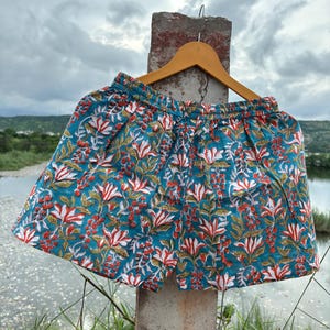 K&ouml;nnte beinhalten: T&uuml;rkise Baumwollshorts mit einem Blumendruck mit wei&szlig;en und roten Bl&uuml;ten. Die Shorts haben einen elastischen Bund und einen Kordelzug.