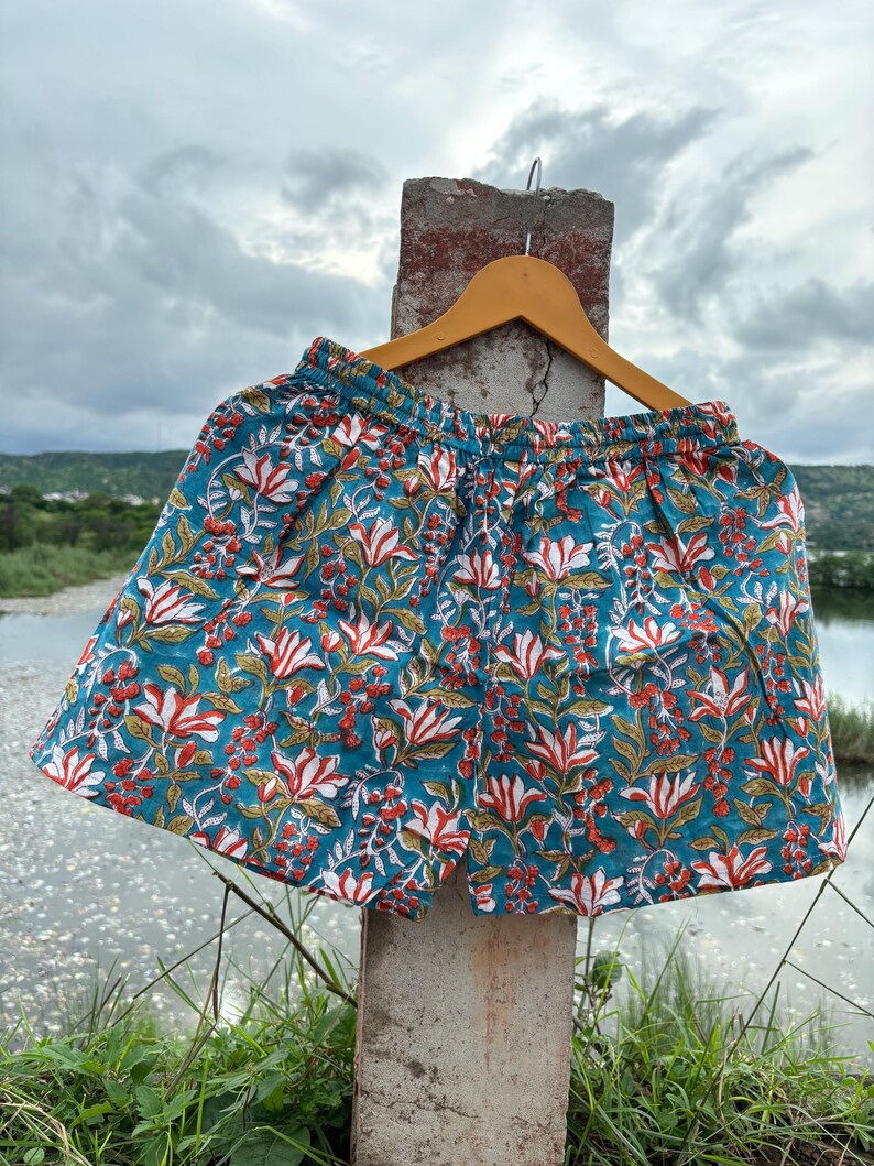 K&ouml;nnte beinhalten: T&uuml;rkise Baumwollshorts mit einem Blumendruck mit wei&szlig;en und roten Bl&uuml;ten. Die Shorts haben einen elastischen Bund und einen Kordelzug.