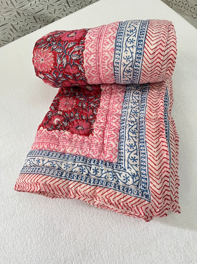 May include: A red and pink cotton quilt with a floral and geometric print. The quilt is folded and rolled up, showing the different patterns on the fabric.