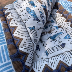 May include: A white and blue quilted throw blanket with a pattern of fish and geometric designs. The blanket is folded in half and has a brown border.