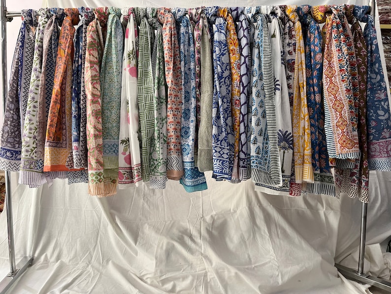 May include: A collection of colorful cotton scarves with various floral and geometric patterns hanging on a rack. The scarves are all different colors and designs, and they are all made of cotton.