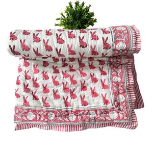 May include: White cotton quilt with a pink bunny rabbit print and a pink and white floral border. The quilt is folded in half and has a green plant in the background.