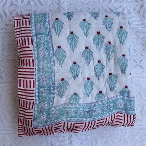 May include: A white quilt with a repeating pattern of teal leaf-like shapes and red accents. The border features a red and white striped design and a teal floral pattern. The quilt is folded and displayed on a white patterned surface.