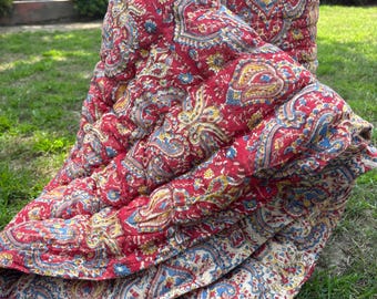May include: A folded red and cream paisley patterned quilt. The quilt features a detailed floral design in shades of red, blue, and yellow. The quilt is lying on green grass.