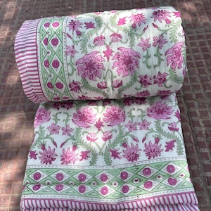 May include: A folded white quilt with a floral pattern in pink and green. The quilt features large pink flowers and green vines on a white background. The border has a striped design in pink and white.