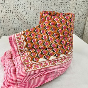 May include: A pink and orange floral patterned quilt with a white background. The quilt is folded in half and has a pink and white striped border.