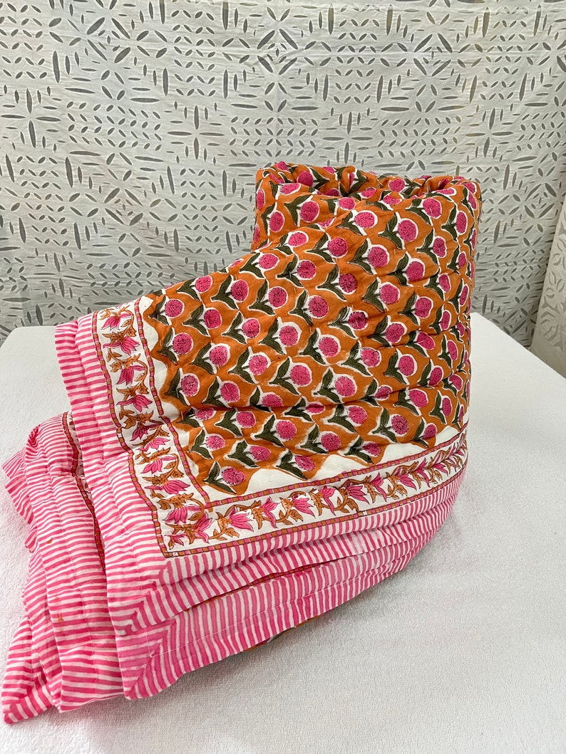 May include: A pink and orange floral patterned quilt with a white background. The quilt is folded in half and has a pink and white striped border.