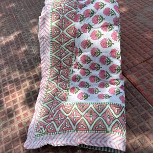May include: A folded quilt with a floral pattern. The quilt has a white background with pink flower motifs. The border features a pink and green geometric design. The quilt is folded on a red surface.