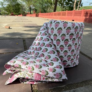 Puede incluir: Un edredón blanco con un patrón repetitivo de diseños florales rosas y hojas verdes. El edredón está parcialmente enrollado y parcialmente doblado, mostrando el intrincado patrón. La ropa de cama está colocada sobre una superficie de piedra.
