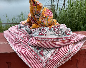 May include: A yellow and pink floral patterned quilt with white stripes and a green border. The quilt is folded and draped over a red brick wall.