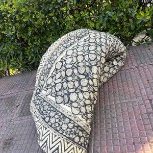Puede incluir: Una colcha plegada con un diseño floral en blanco y negro. La colcha tiene un borde con un patrón geométrico en zigzag. El fondo presenta follaje verde y una superficie de baldosas marrón rojizo.