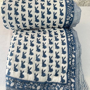 May include: A white cotton quilt with a blue bunny print and a blue floral border. The quilt is folded in half and the border is visible on both sides.