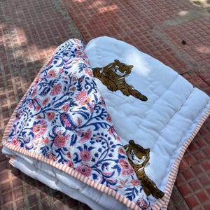 May include: A folded white and floral patterned quilt with a tiger print. The quilt features a white background with brown tigers and a blue, pink, and white floral pattern. The quilt is trimmed with a peach and white striped border.