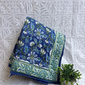 May include: A folded blue quilt with a floral pattern in white and green. The quilt has a border with a similar design. The background is a white fabric with a repeating pattern. A small potted plant is in the lower right corner.
