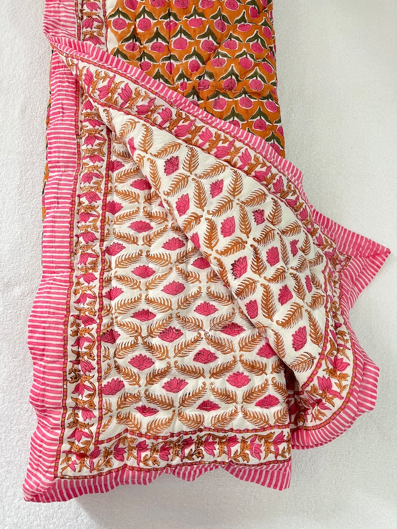 May include: A white cotton quilt with a pink and orange floral print. The quilt is folded in half and has a pink and white striped border.