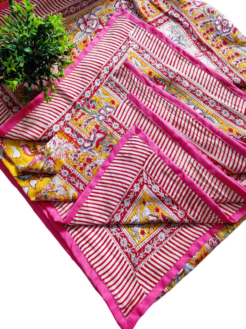 May include: A set of four pink and white striped placemats with a yellow floral pattern and pink trim. The placemats are folded and stacked on top of each other.