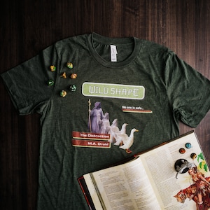 Op de afbeelding: Een donkergroen T-shirt met een afbeelding van een druïde in een bosrijke omgeving. De afbeelding bevat de tekst "Wild Shape" en "The Distraction M.A. Druid".