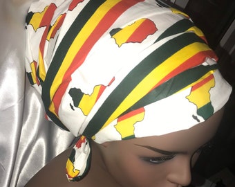 Puede incluir: Un turbante blanco con un patrón de rayas amarillas, rojas y verdes y contornos del continente africano. El turbante está atado en un nudo en la parte posterior de la cabeza.