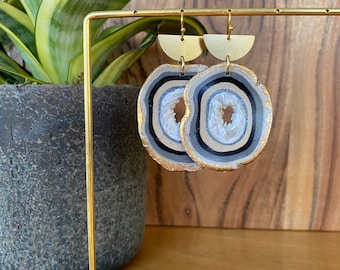 May include: A pair of agate-style earrings with gold-colored accents. The earrings feature a layered design with shades of gray, black, and white, and are suspended from gold-colored half-circle tops. The earrings are displayed on a gold-colored stand.