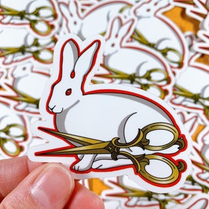 May include: A white rabbit sticker with red accents and gold scissors. The sticker has a red outline and a drop of red on the rabbit's head. The scissors are positioned across the rabbit's body.