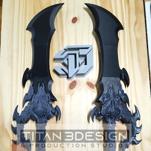 May include: Two black and grey fantasy swords with detailed handles and curved blades. A grey logo is above the swords. The text "TITAN 3DESIGN & PRODUCTION STUDIOS" is printed below the swords.